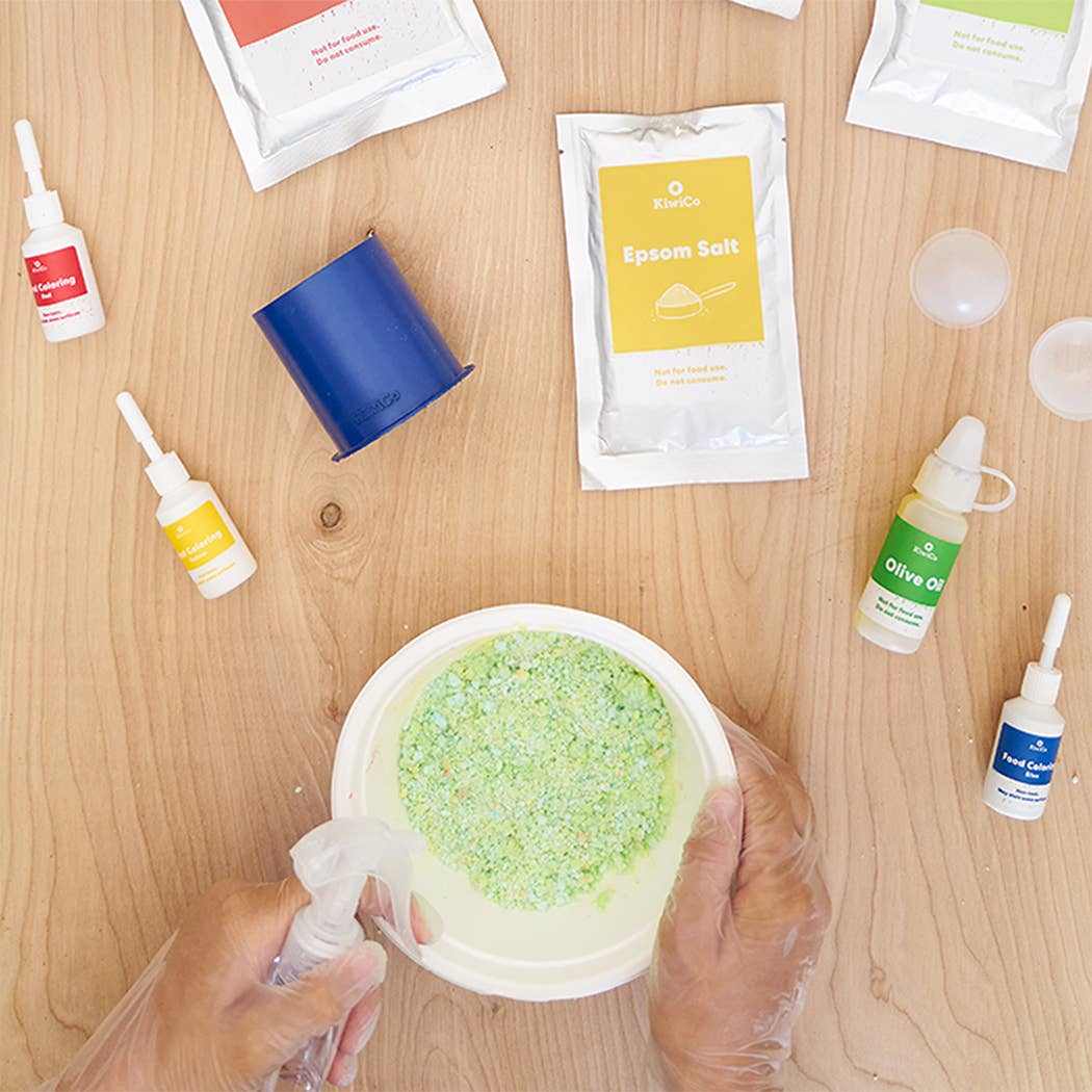 Hands mixing green bath bomb powder in a bowl surrounded by Planet Bath-Bombs Science Kit ingredients and tools on wooden table
