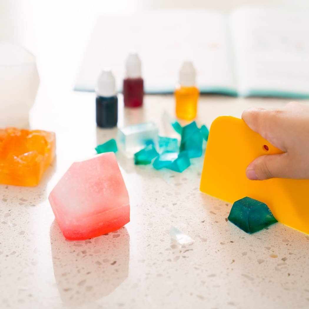 Child using Crystal Geode Soapmaking Kit to cut colorful crystal-inspired soaps on a countertop with liquid colors in the background