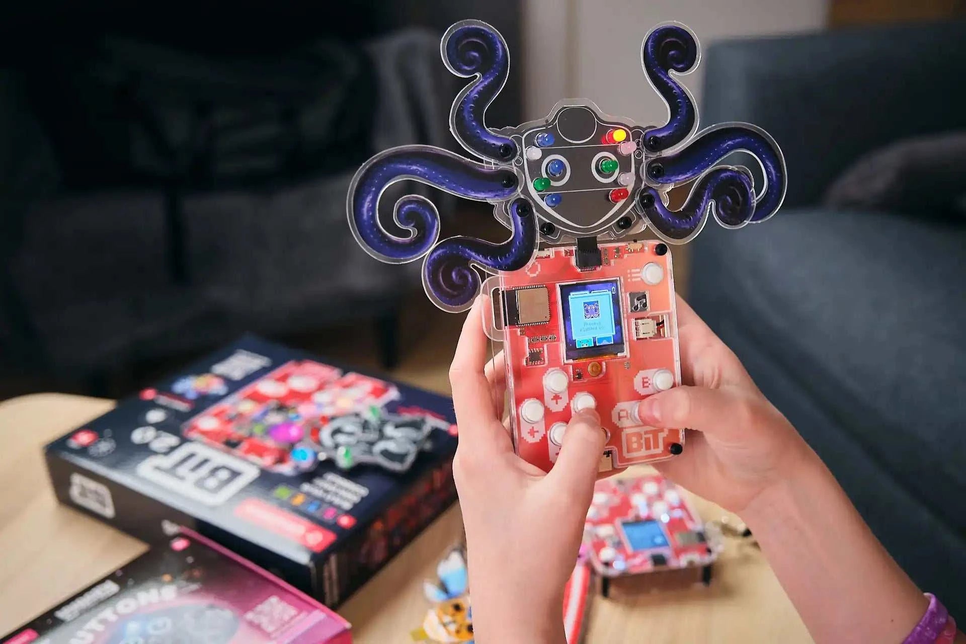 Hands holding Wacky Robot Artemis mini robot with lit LEDs and game screen, circuit board and octopus design in focus on table with packaging in background