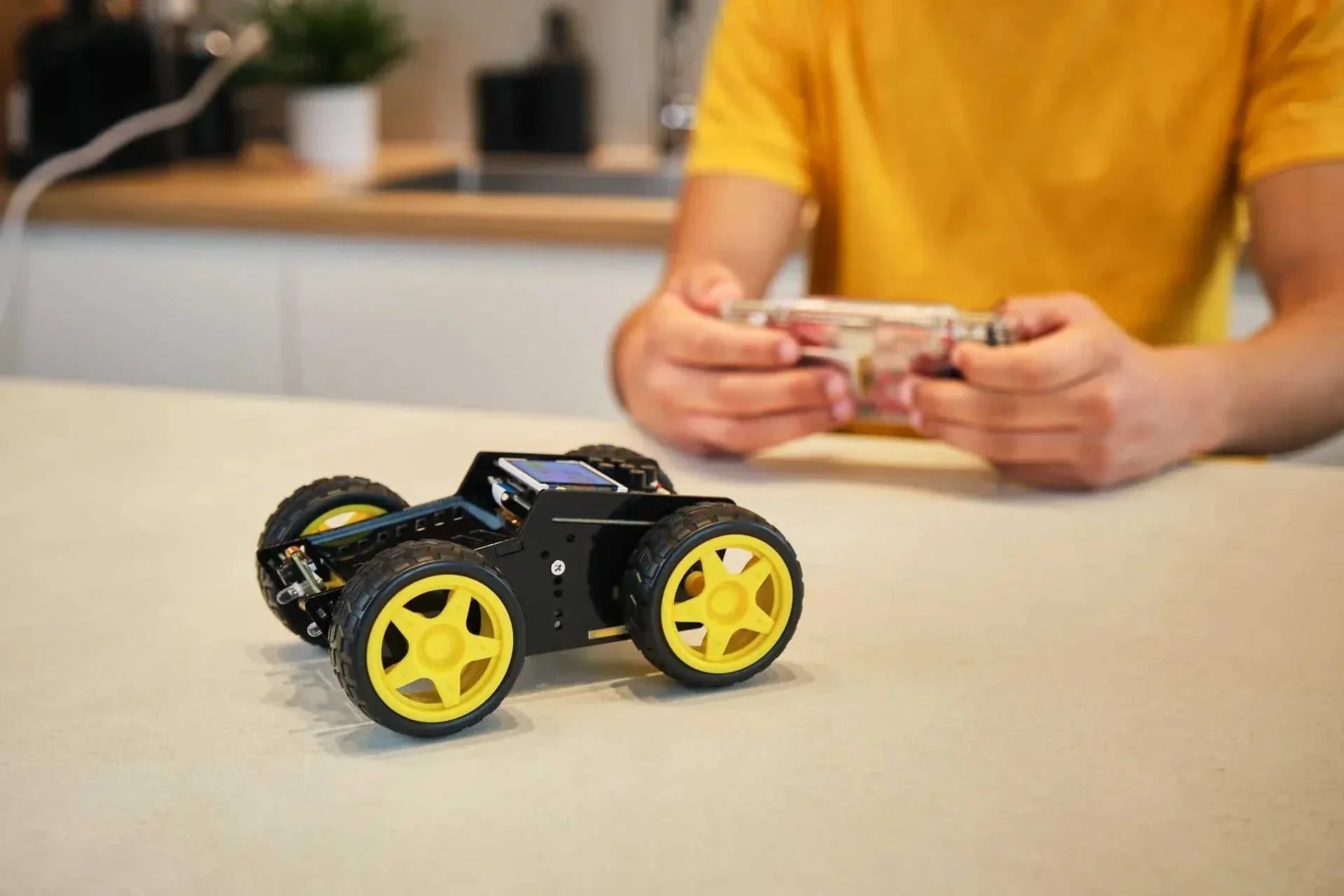 Wheelson AI-powered self-driving robot car with yellow wheels on a table and person controlling it with a remote in the background