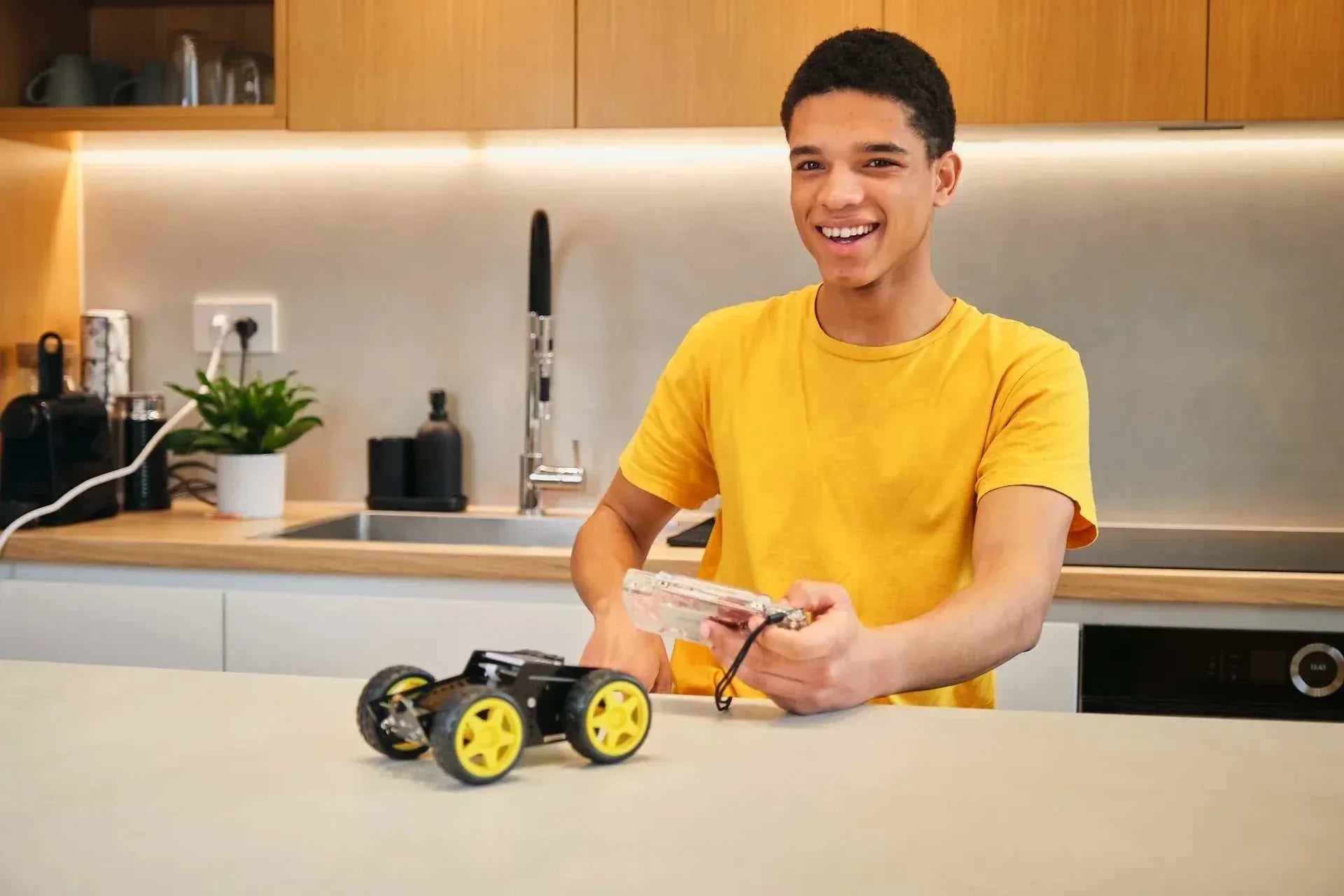Teen boy in yellow shirt assembling and testing Wheelson AI-powered self-driving robot car with yellow wheels on kitchen counter
