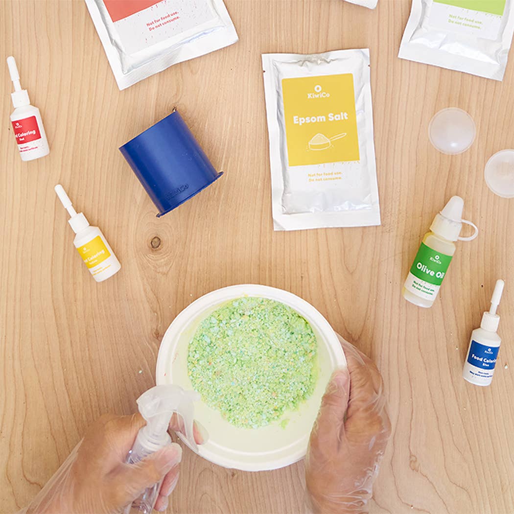 Hands mixing green bath bomb powder in a bowl surrounded by Planet Bath-Bombs Science Kit ingredients and tools on wooden table