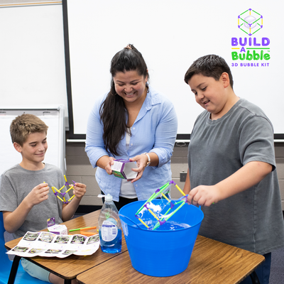 Children using the Build-A-Bubble Montessori 3D bubble kit to create geometric bubble shapes with adult supervision at a table with soap solution and instruction booklet.